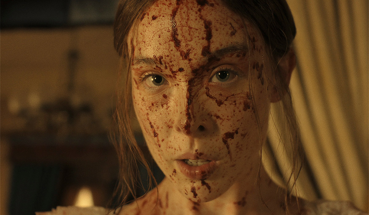 "A young woman with long hair, dressed in light clothing, has her face and upper chest covered in blood, standing indoors with dim warm lighting and blurred curtains and furnishings in the background."